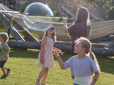 Mehrere Kinder spielen bei Sonnenschein im Gartenbereich des Hotels Unterschwarzachhof mit großen Seifenblasen, die von einer Betreuerin mit einer Schlaufe erzeugt werden.