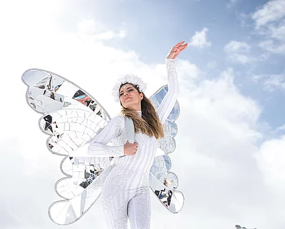 Eine Frau in glitzerndem weißen Kostüm mit verspiegelten Schmetterlingsflügeln und Blumenschmuck posiert vor alpiner Kulisse und strahlend blauem Himmel in Hinterglemm.
