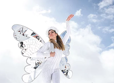 Eine Frau in glitzerndem weißen Kostüm mit verspiegelten Schmetterlingsflügeln und Blumenschmuck posiert vor alpiner Kulisse und strahlend blauem Himmel in Hinterglemm.