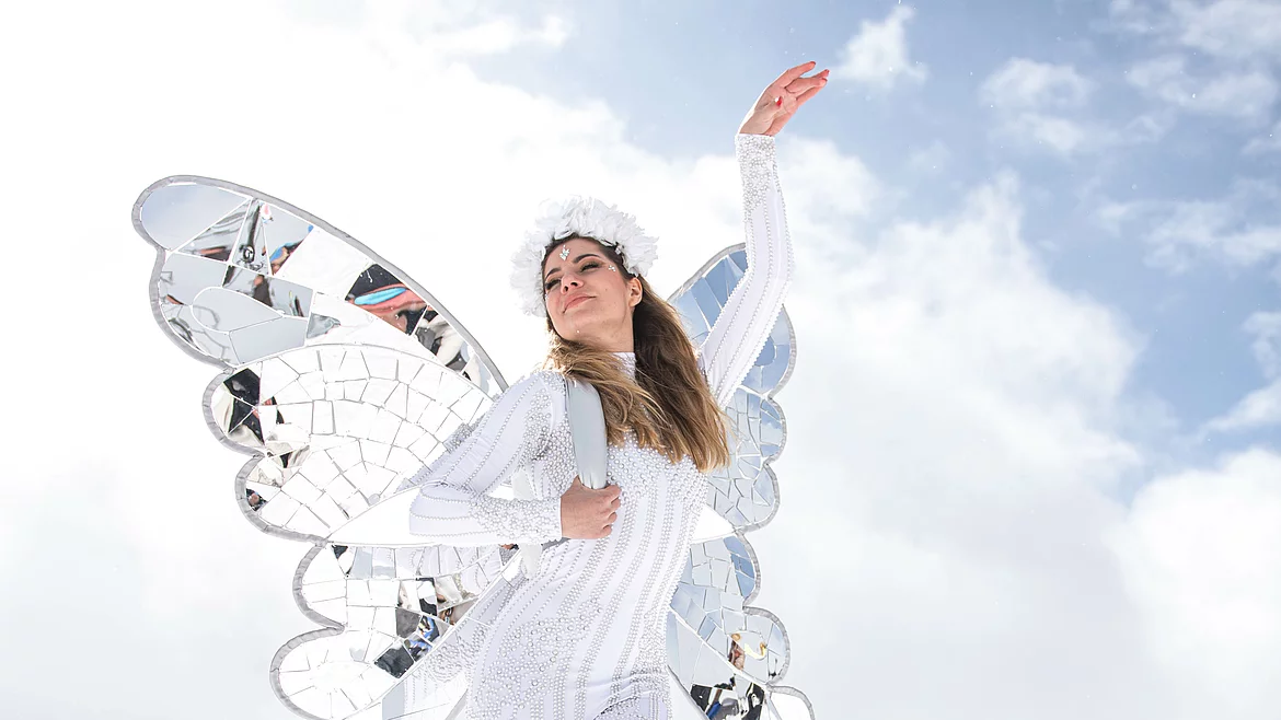 Eine Frau in glitzerndem weißen Kostüm mit verspiegelten Schmetterlingsflügeln und Blumenschmuck posiert vor alpiner Kulisse und strahlend blauem Himmel in Hinterglemm.