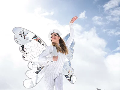 Eine Frau in glitzerndem weißen Kostüm mit verspiegelten Schmetterlingsflügeln und Blumenschmuck posiert vor alpiner Kulisse und strahlend blauem Himmel in Hinterglemm.