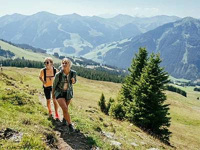 Zwei Frauen wandern bei strahlendem Sonnenschein auf einem alpinen Wanderweg in den Bergen rund um Hinterglemm, mit weitem Blick ins grüne Tal und auf umliegende Gipfel.