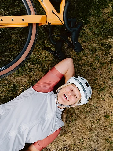 Radfahrerin in Sportkleidung liegt lachend mit Gravelbike auf einer Wiese