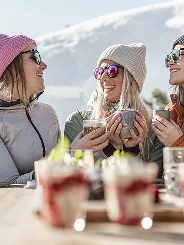 Drei Frauen trinken heiße Getränke auf Sonnenterrasse im Skigebiet mit Winterkulisse